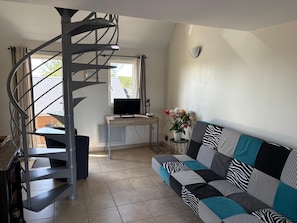 A modern living room with a spiral staircase, a flat-screen TV, a sofa with patterned cushions, and a small side table with a vase of flowers. - Charming Normandy Escape for Three (Courtils)