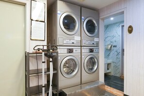A laundry facility with four stacked washers and dryers, a metal shelf, and a door leading to a bathroom. - Paco Hotel (Guangzhou Tianhe Coach Terminal Metro Station) (Guangzhou)
