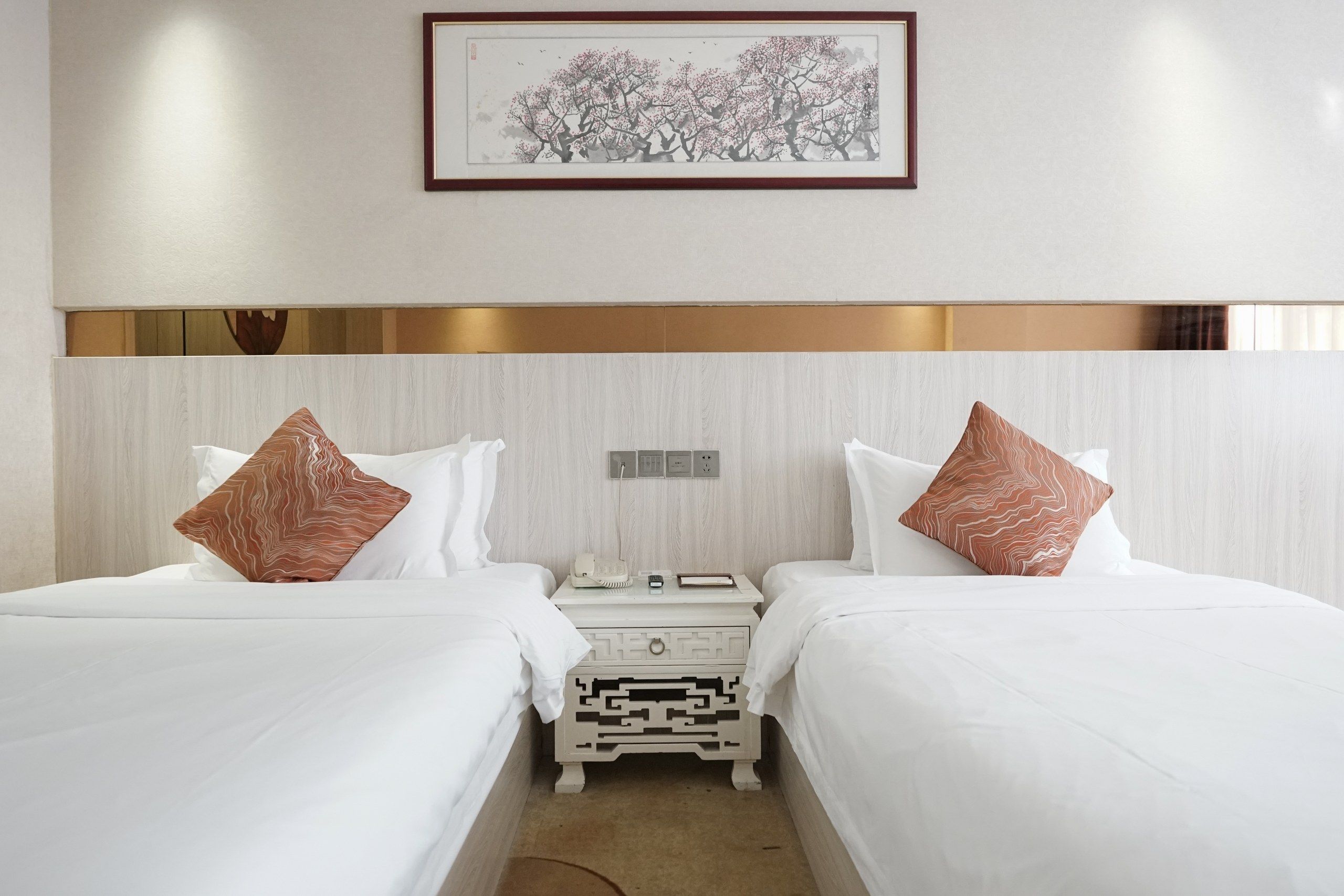 A hotel room with two beds, a bedside table with a phone and a lamp, and a framed artwork on the wall.