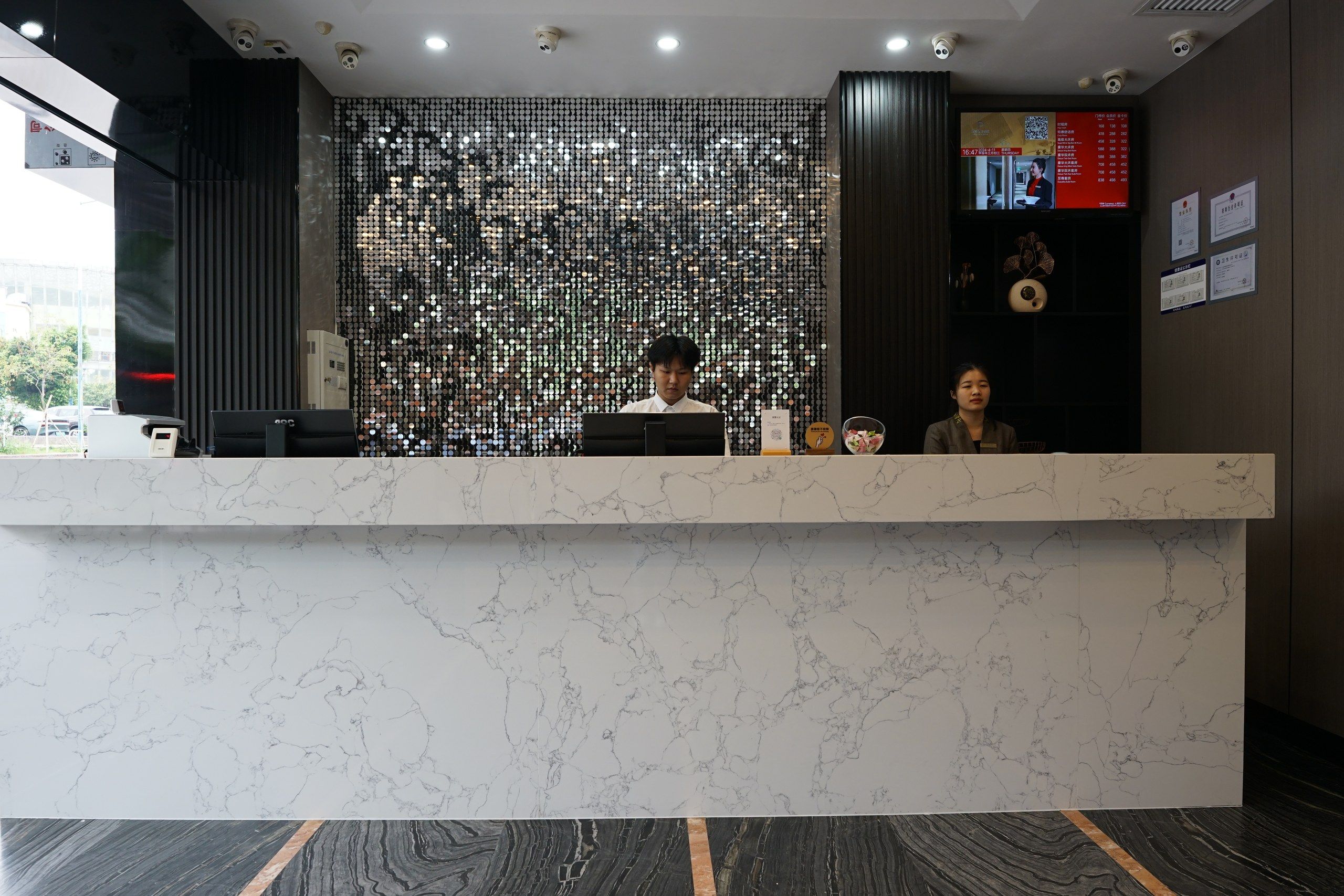 Un mostrador de recepción con encimera de mármol, una pantalla digital que muestra información y una pared decorativa con patrones circulares.