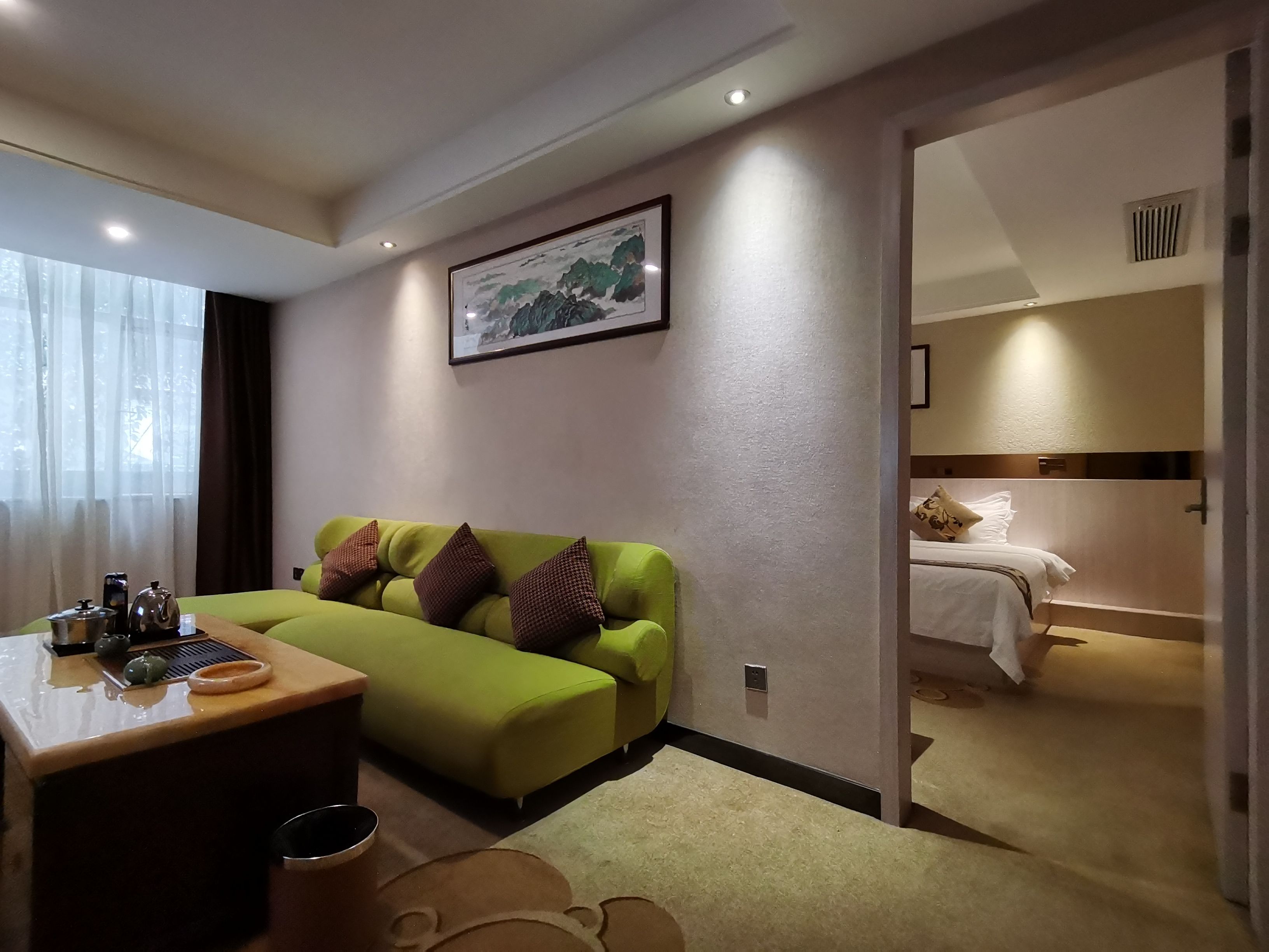 Quarto de hotel com sofá verde, uma pequena mesa com conjunto de chá, um quadro na parede e uma cama com cabeceira.