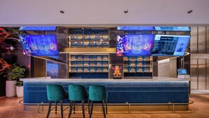 Uma área de bar moderna com bancada de mármore, banquetas verdes e várias telas digitais exibindo conteúdo.