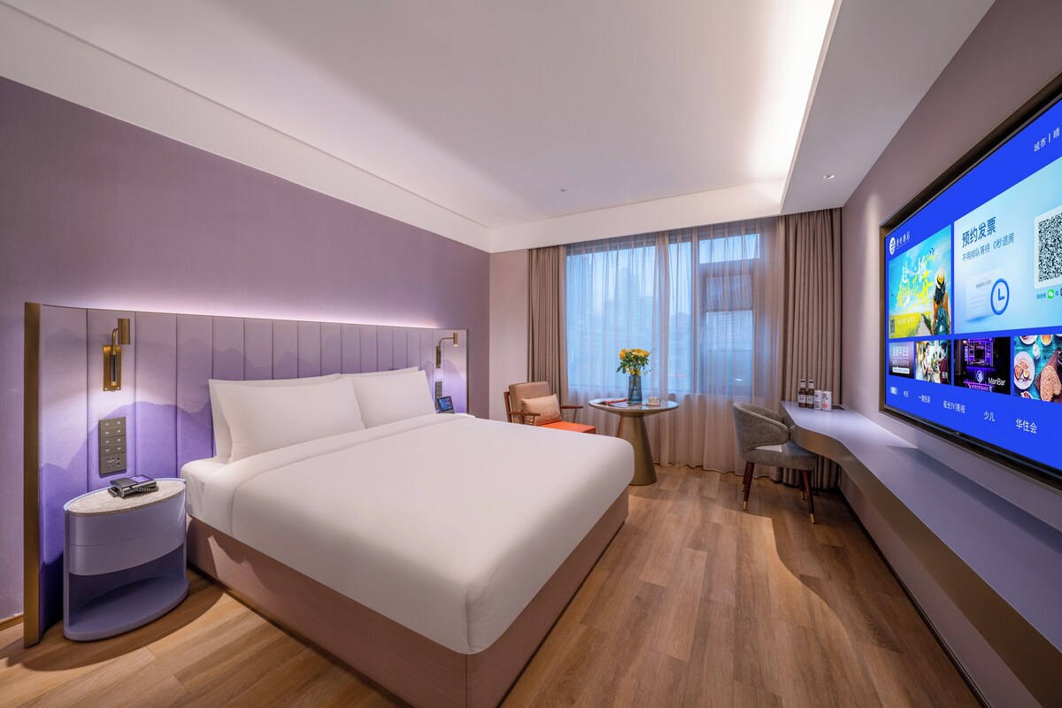A modern hotel room with a large bed, bedside tables, a desk, and a TV displaying a digital interface.