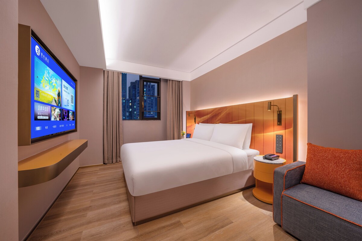 A hotel room with a large bed, a TV mounted on the wall, a window with curtains, a bedside table, and a small seating area with a cushion.
