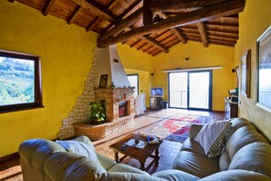 A living room with a stone fireplace, a sofa, a coffee table, and a large window. - Villa Elsa, with a pool and breathtaking views of the Roman countryside (Lazio)