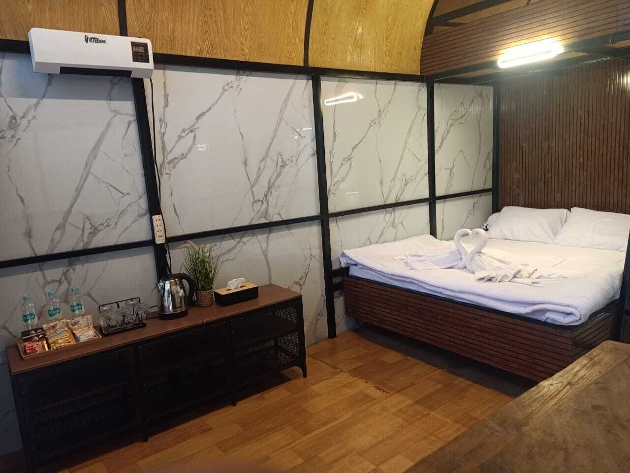 A hotel room with a bed, a wooden bedside table with amenities, a marble-patterned wall, and a ceiling-mounted air conditioning unit.