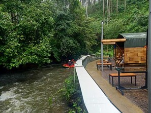 Un paseo junto al río con un puente de madera, bancos y una zona de estar cubierta.
