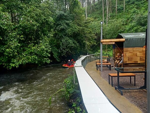Un paseo junto al río con un puente de madera, bancos y una zona de estar cubierta.