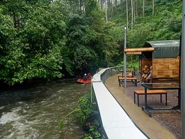 Un paseo junto al río con un puente de madera, bancos y una zona de estar cubierta.