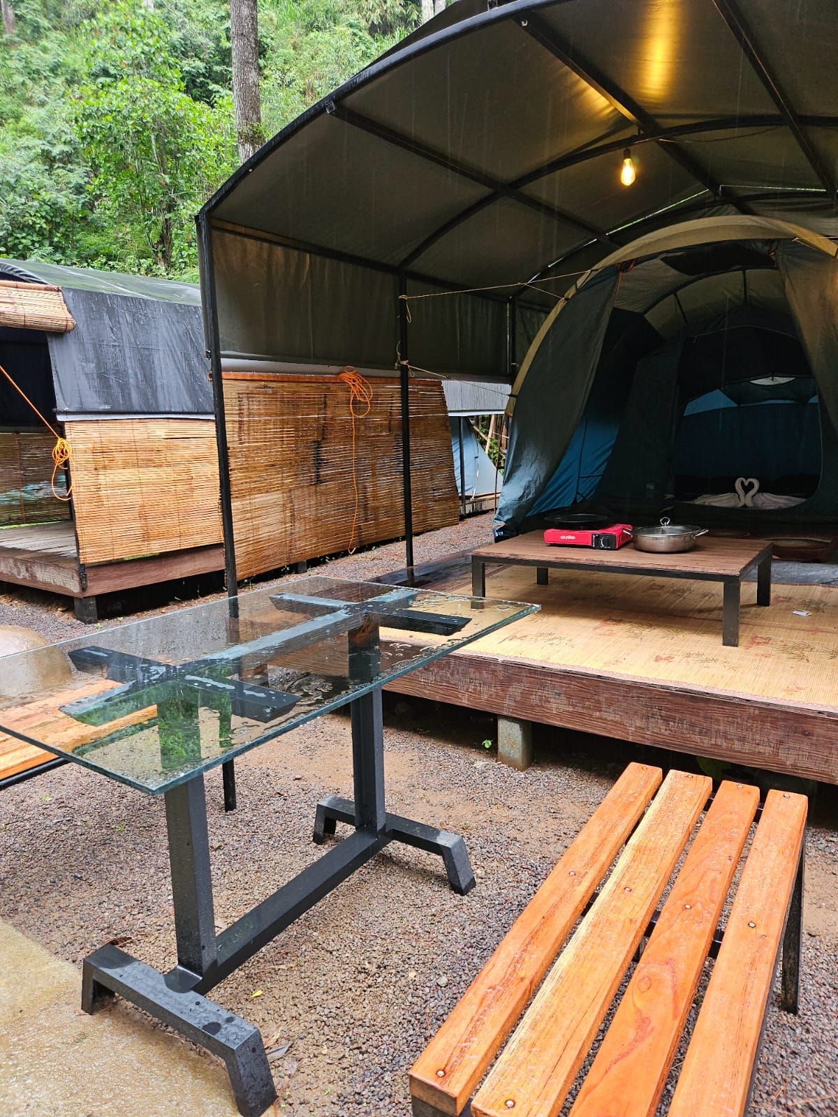 Un espace de camping aménagé avec une zone de repas couverte, une table en verre, des bancs en bois et une tente.