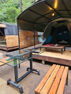 Un espace de camping aménagé avec une zone de repas couverte, une table en verre, des bancs en bois et une tente.