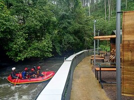 Un río con una balsa roja y personas con cascos. Hay una pasarela con bancos y una zona cubierta con mesas.