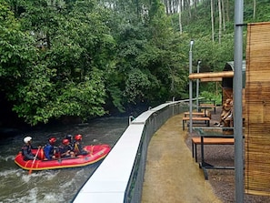 A river with a red raft and people wearing helmets. There is a walkway with benches and a covered area with tables. - Cozzy Haruman riverside camp (Pangalengan)