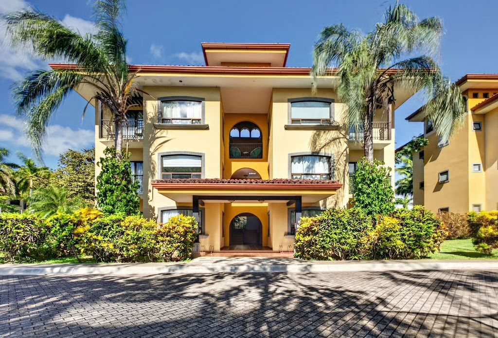 A multi-story hotel with a central entrance, balconies, and surrounding palm trees.