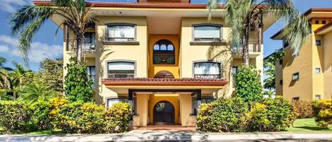 A multi-story hotel with a central entrance, balconies, and surrounding palm trees.