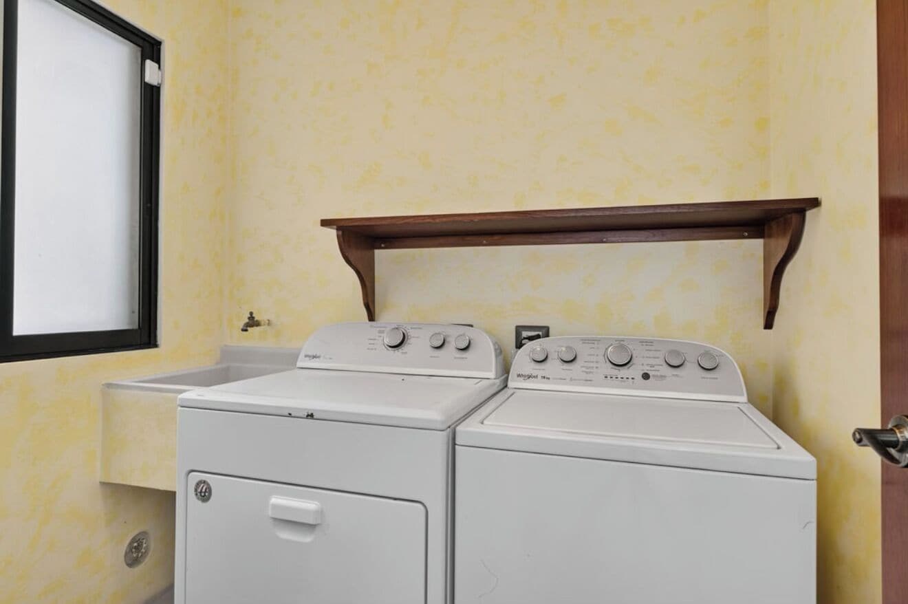 A laundry room with two front-loading washing machines and a wooden shelf above them.