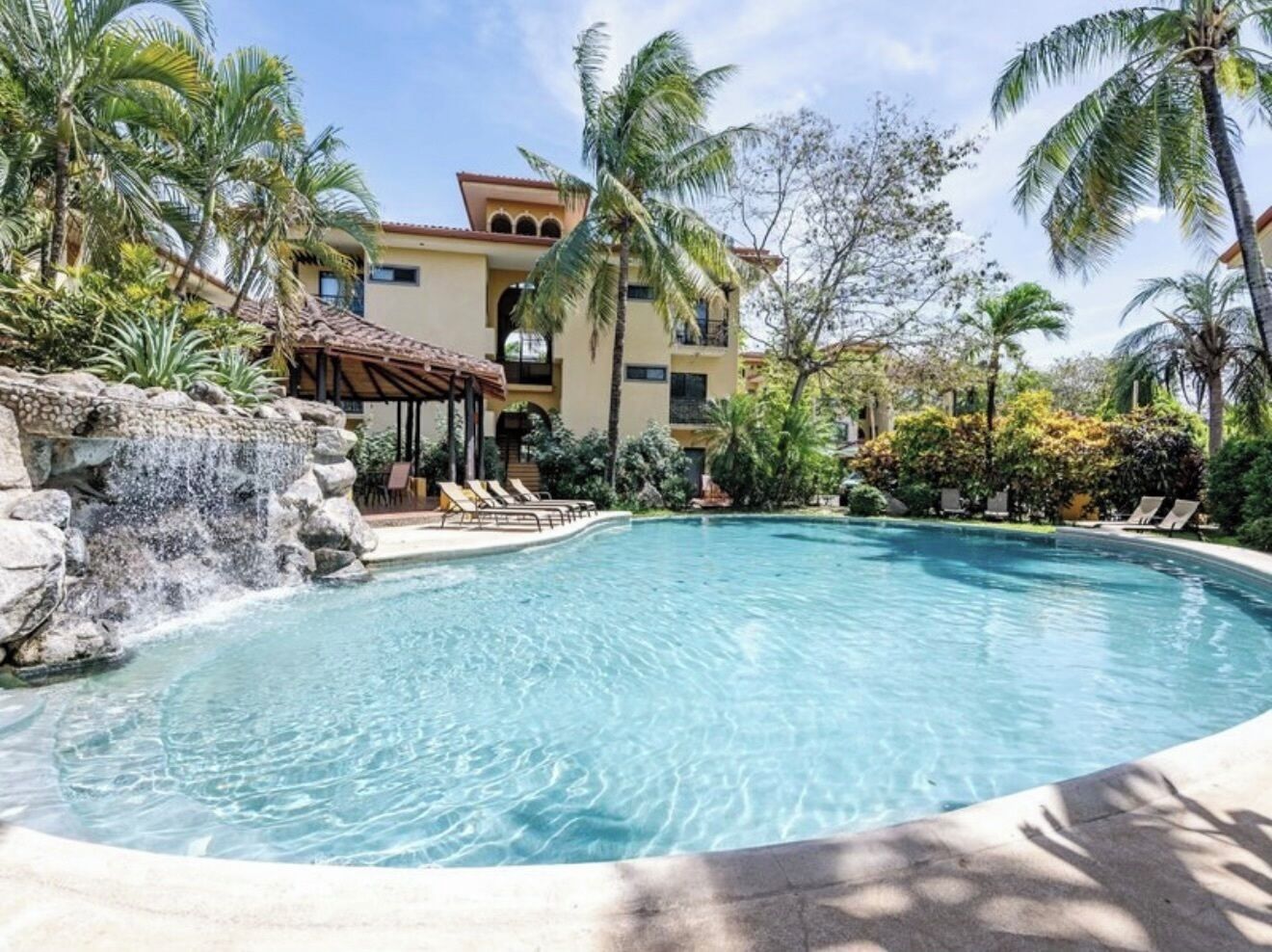 A swimming pool with a waterfall feature, surrounded by palm trees and lounge chairs.