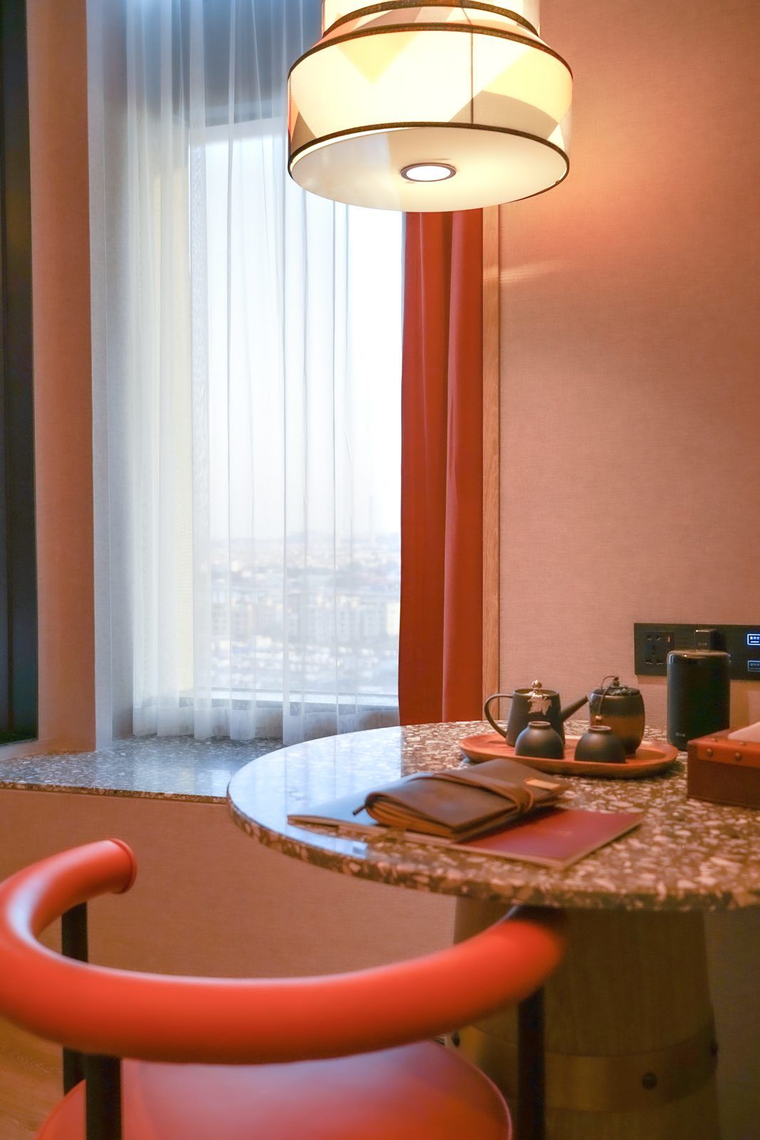 A hotel room with a round table, a red chair, a tea set, and a view through a window with sheer curtains.