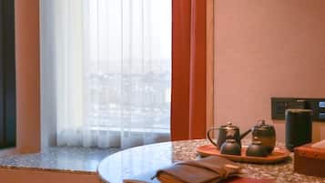 A hotel room with a round table, a red chair, a tea set, and a view through a window with sheer curtains.
