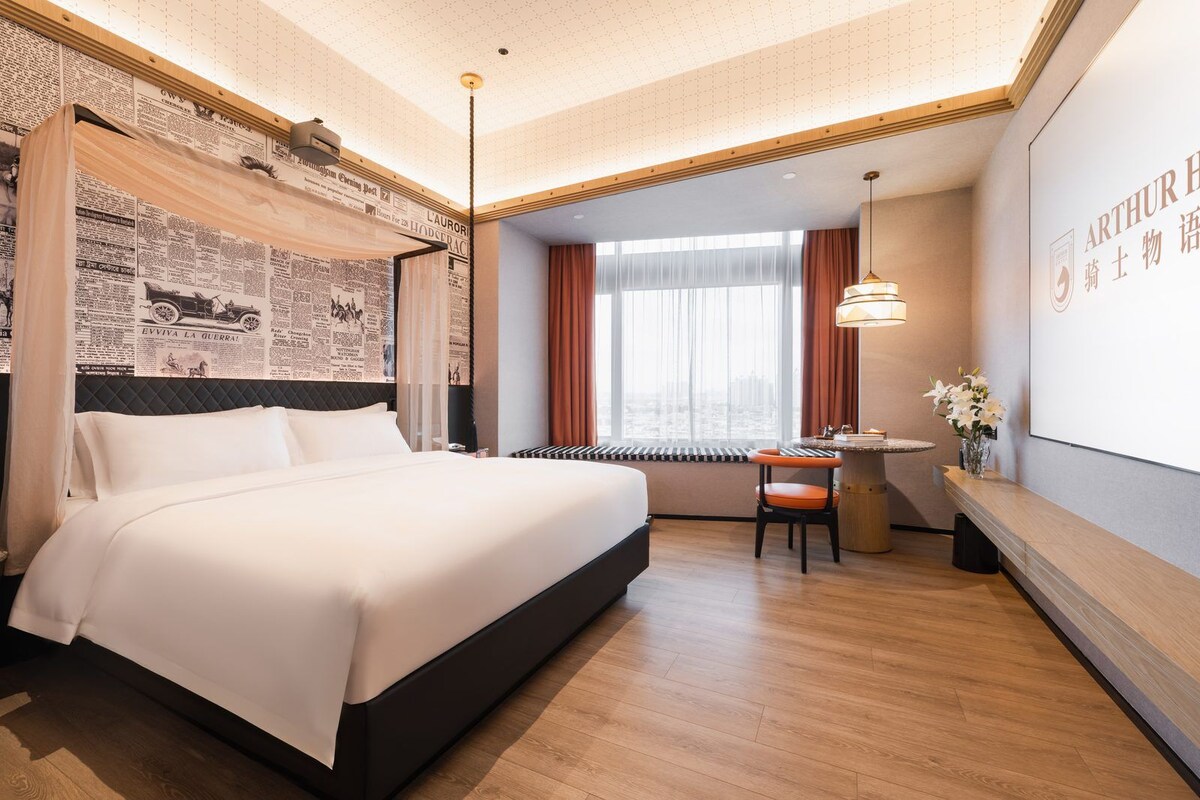Quarto de hotel com uma cama grande, uma escrivaninha com cadeira, uma janela com cortinas e um quadro na parede.