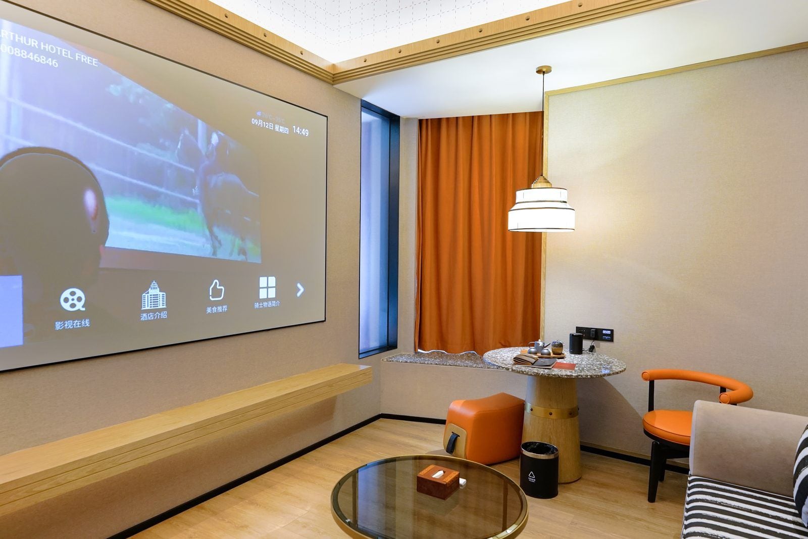 A hotel room with a projection screen displaying a movie, a round table with a lamp, a sofa, and a small trash bin.