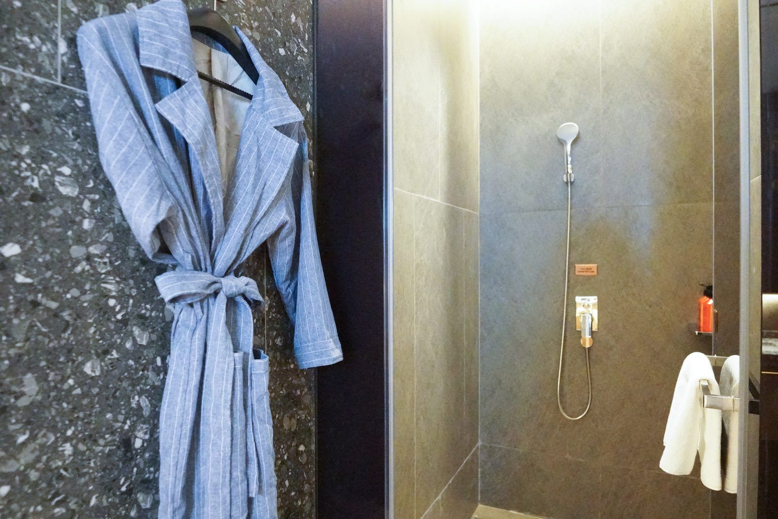 Un baño con piso de mármol, una bata colgada en un perchero, una ducha con cabezal de ducha manual y un toallero con una toalla blanca.