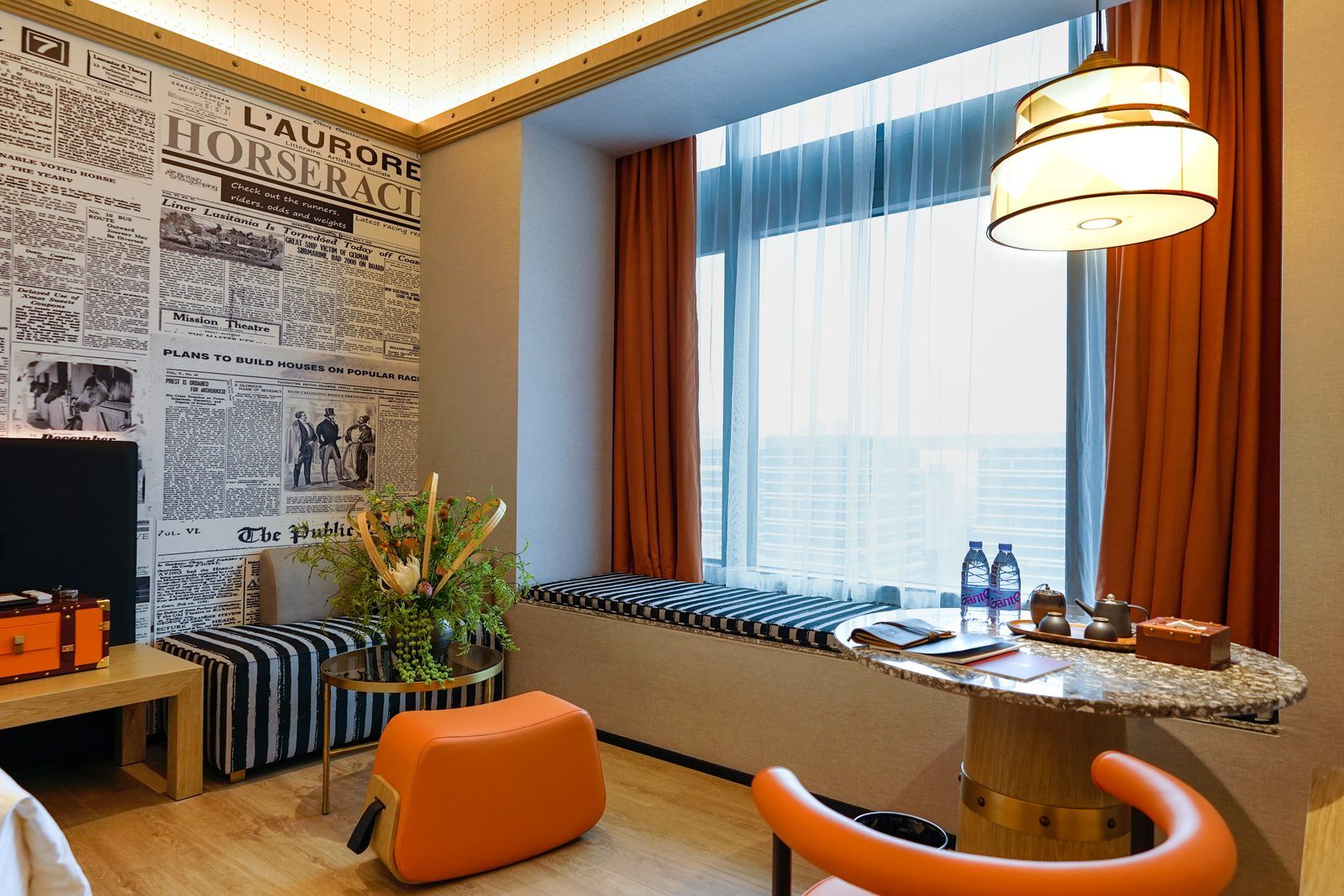 A hotel room with a large window, a bench, a small table with a lamp, and a newspaper on the wall.