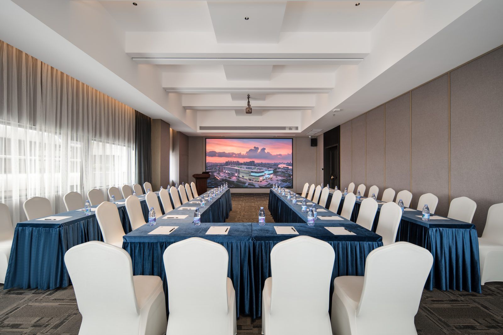 A conference room with a long table, white chairs, and a large screen displaying a cityscape.
