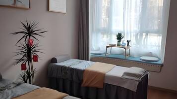 A hotel room with two beds, a large window with curtains, a small table with a plant, and framed pictures on the wall.