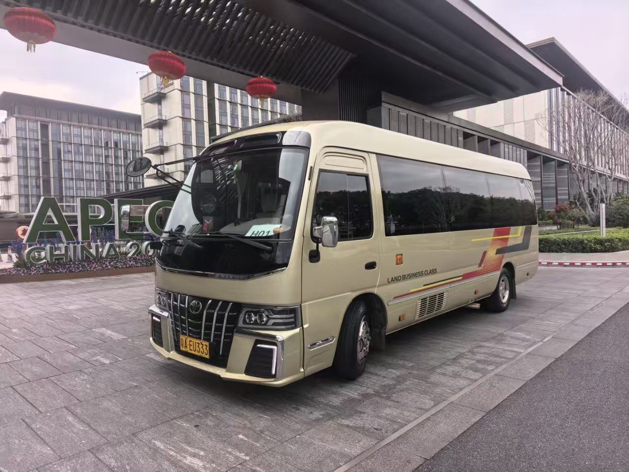 Un autobús color beige con el texto 'LAND BUSINESS CLASS' en la carrocería estacionado frente a un edificio de diseño moderno.