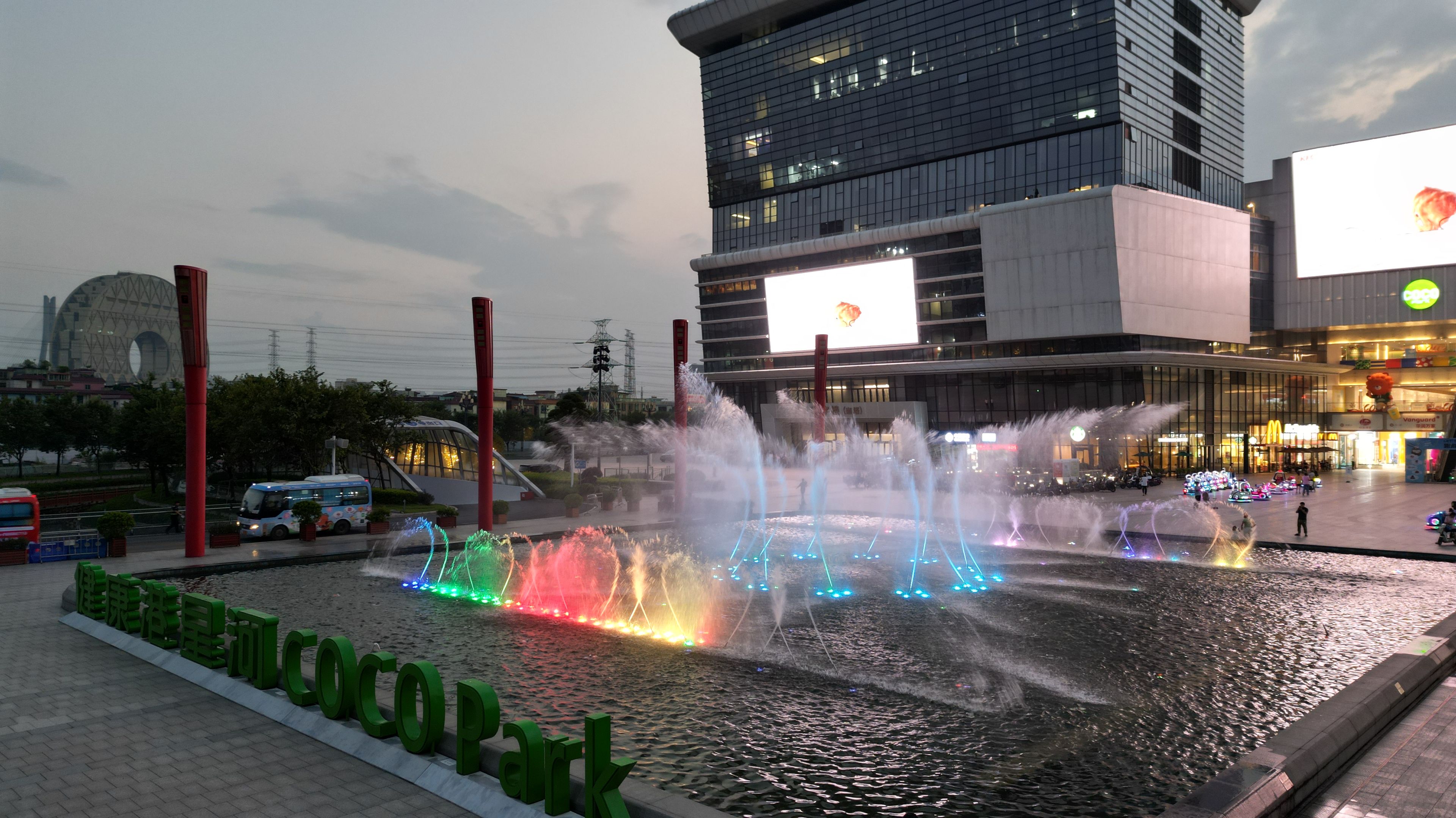 A water fountain with multiple jets and colorful lights in front of a modern building with a large billboard.