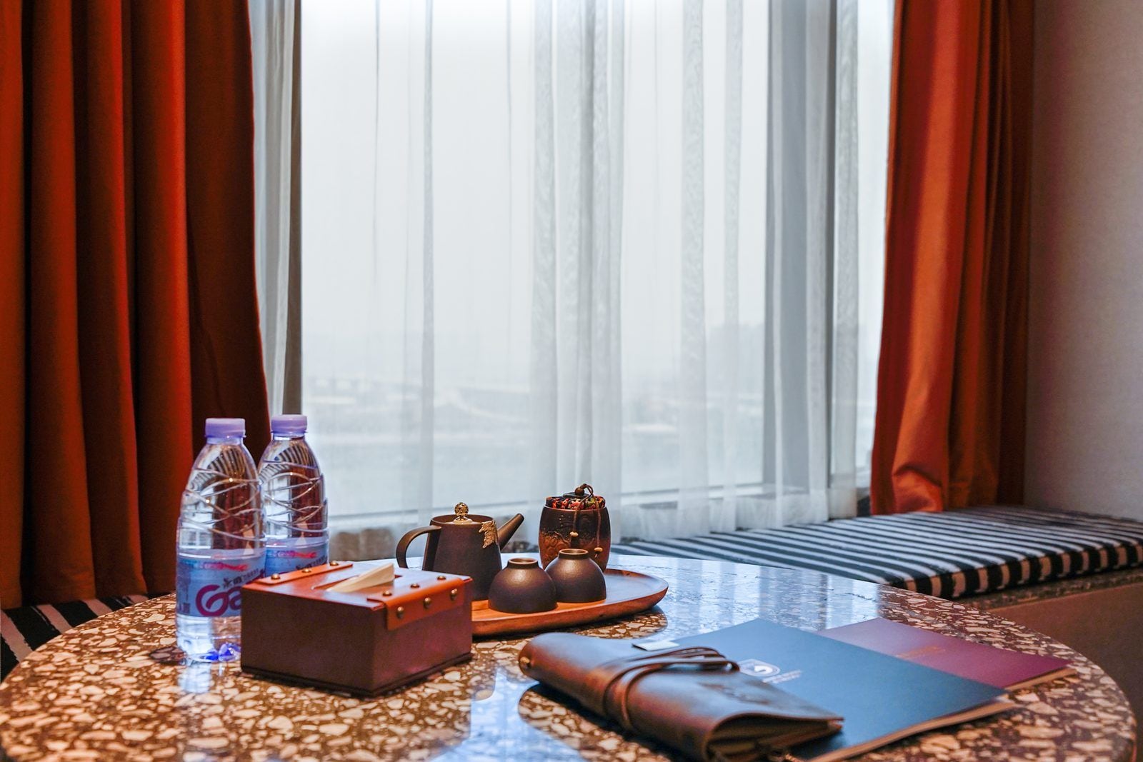 Une chambre d’hôtel avec une table ronde sur laquelle sont disposés une théière, deux bouteilles d’eau, une boîte de mouchoirs et un carnet.