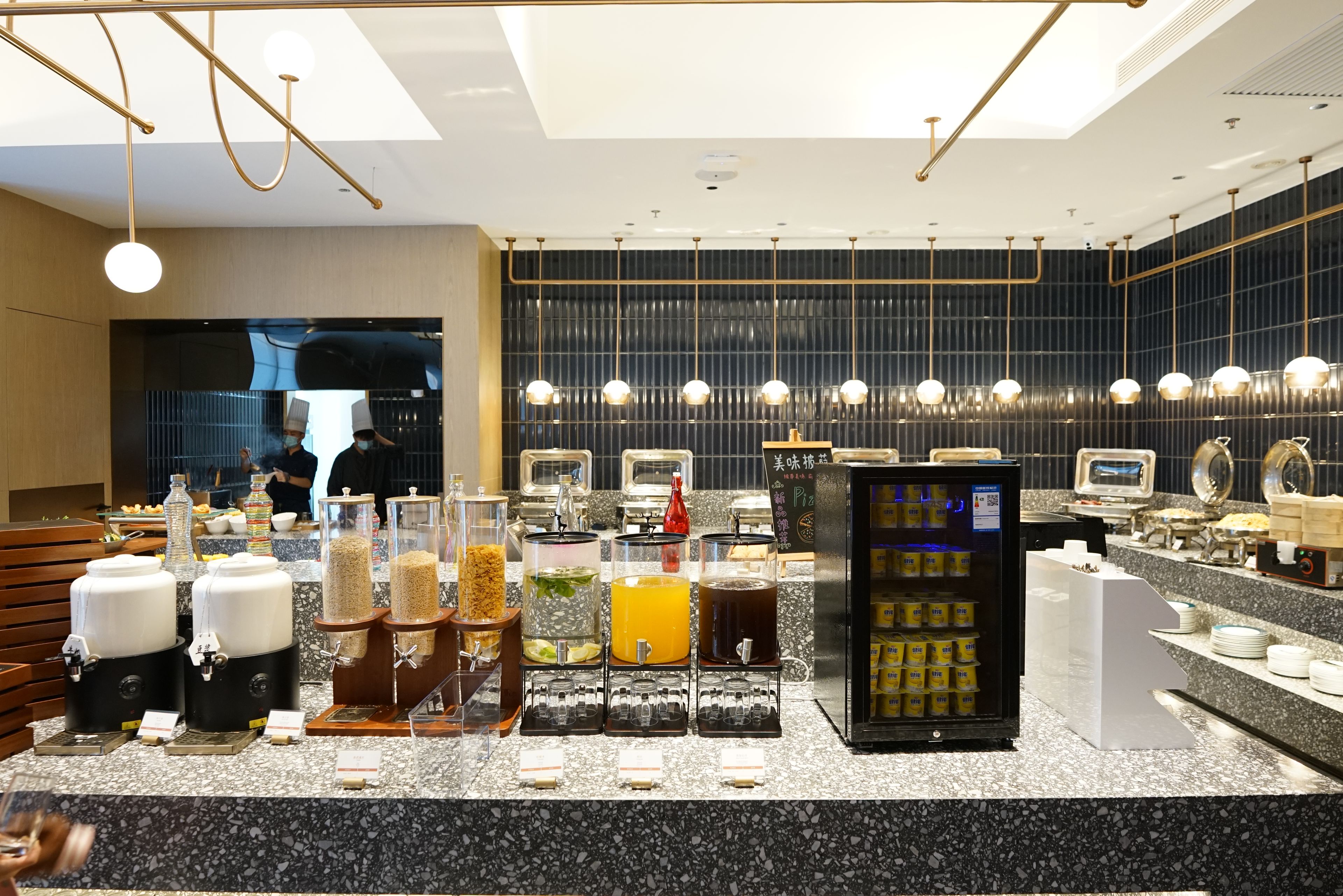 A modern cafe with a self-serve beverage station, a pizza display, and a seating area with wooden benches.