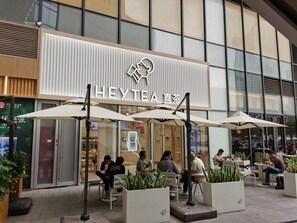 Ett utomhusområde med sittplatser, parasoller och krukväxter framför en byggnad med en skylt som visar texten ”HEYTEA”.