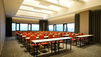 Un salón de conferencias con filas de mesas y sillas, ventanas amplias y un alto techo con luminarias.