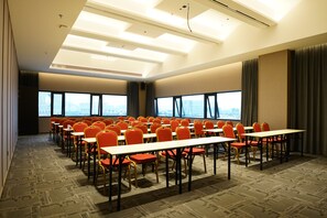 A conference room with rows of tables and chairs, large windows, and a high ceiling with lighting fixtures.