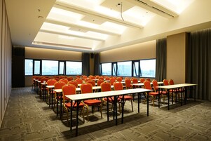 Un salón de conferencias con filas de mesas y sillas, ventanas amplias y un alto techo con luminarias.