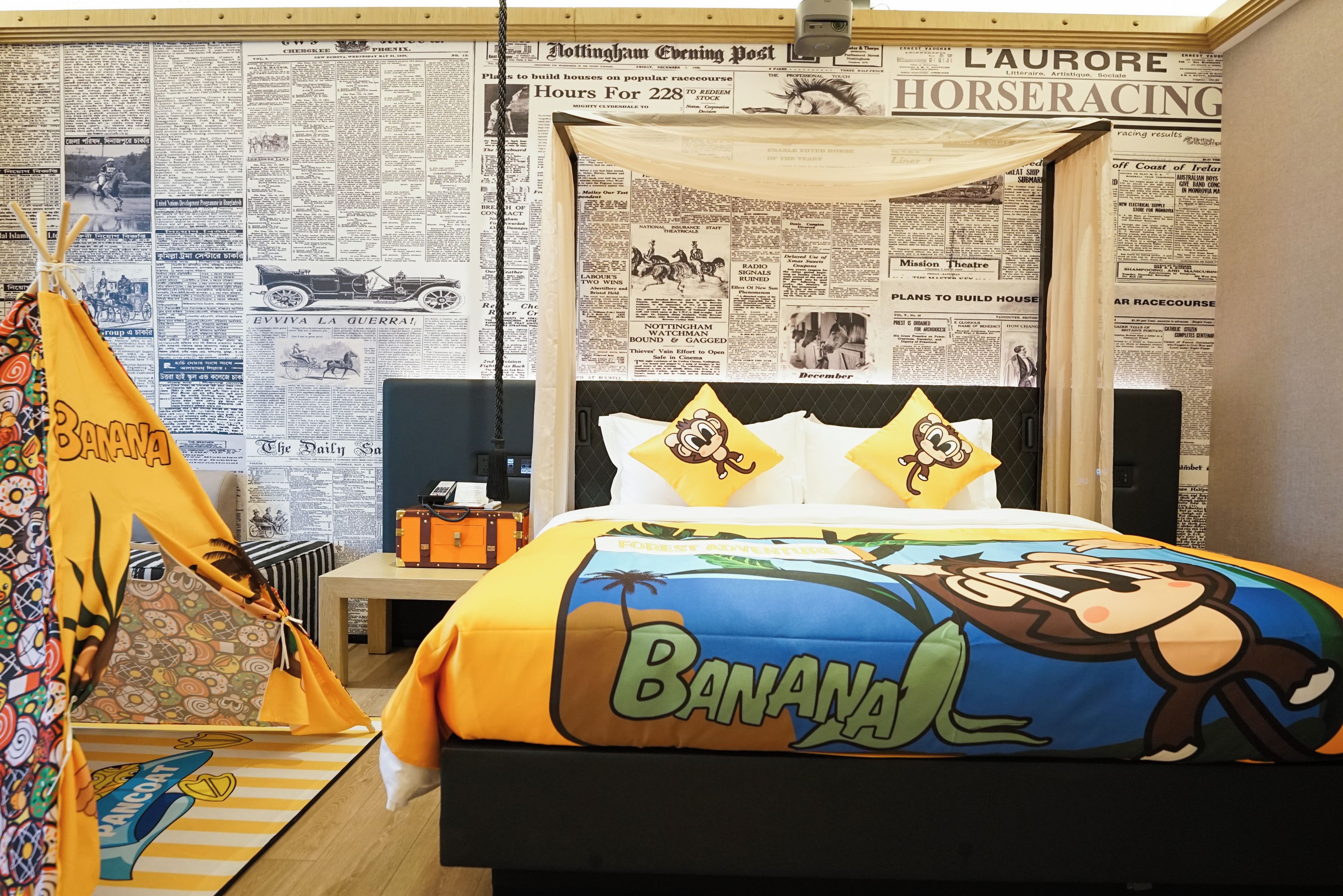 A room with a bed featuring a banana-themed design, a small table with a lamp, and a wall covered with newspaper clippings.