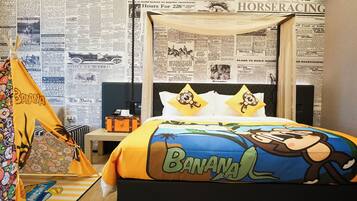 A room with a bed featuring a banana-themed design, a small table with a lamp, and a wall covered with newspaper clippings.