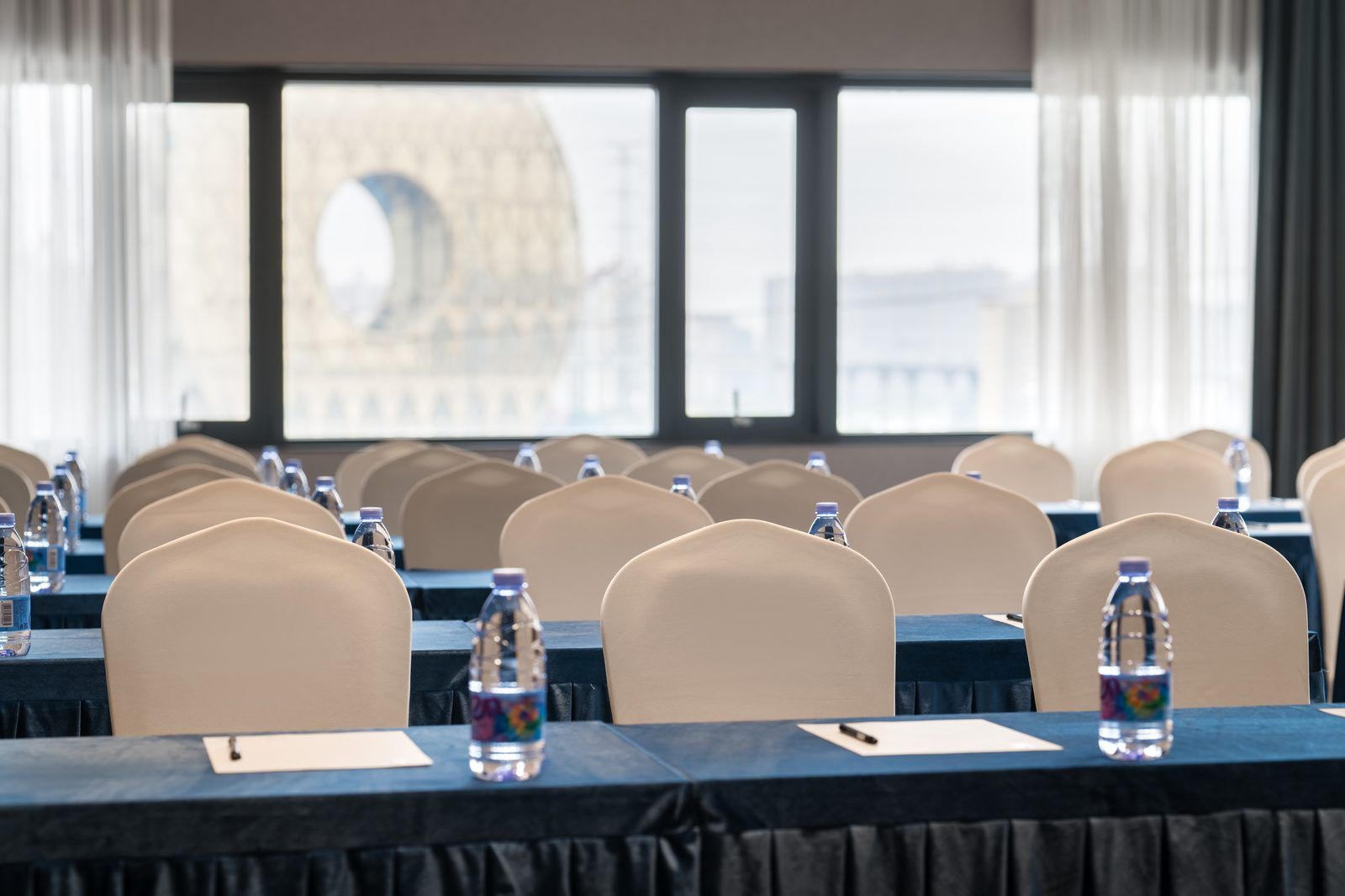 Un salón de conferencias con filas de sillas color beige, mesas con botellas de agua y vista a un edificio grande a través de las ventanas.