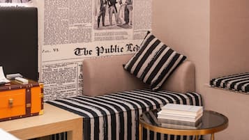 Een kamer met een bank, een gestreept kussen, een klein rond tafeltje met boeken en een wand met kranten.