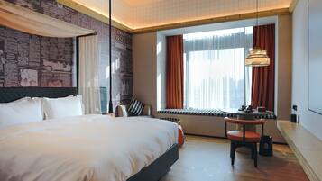 A hotel room with a large bed, a desk with a chair, a window with curtains, and a city view.