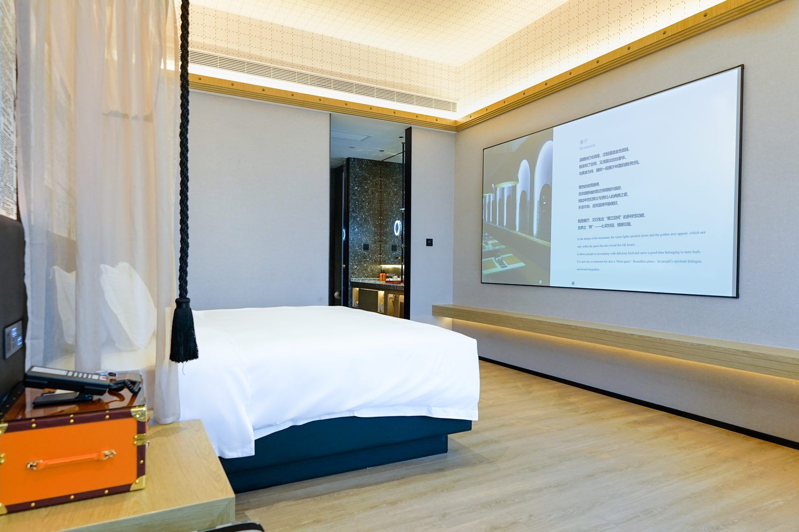 A hotel room with a bed, a desk with a suitcase, and a large screen displaying text.
