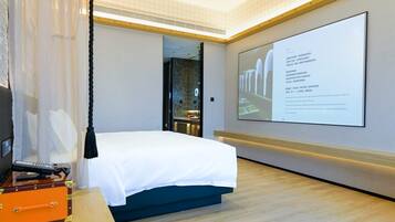 A hotel room with a bed, a desk with a suitcase, and a large screen displaying text.