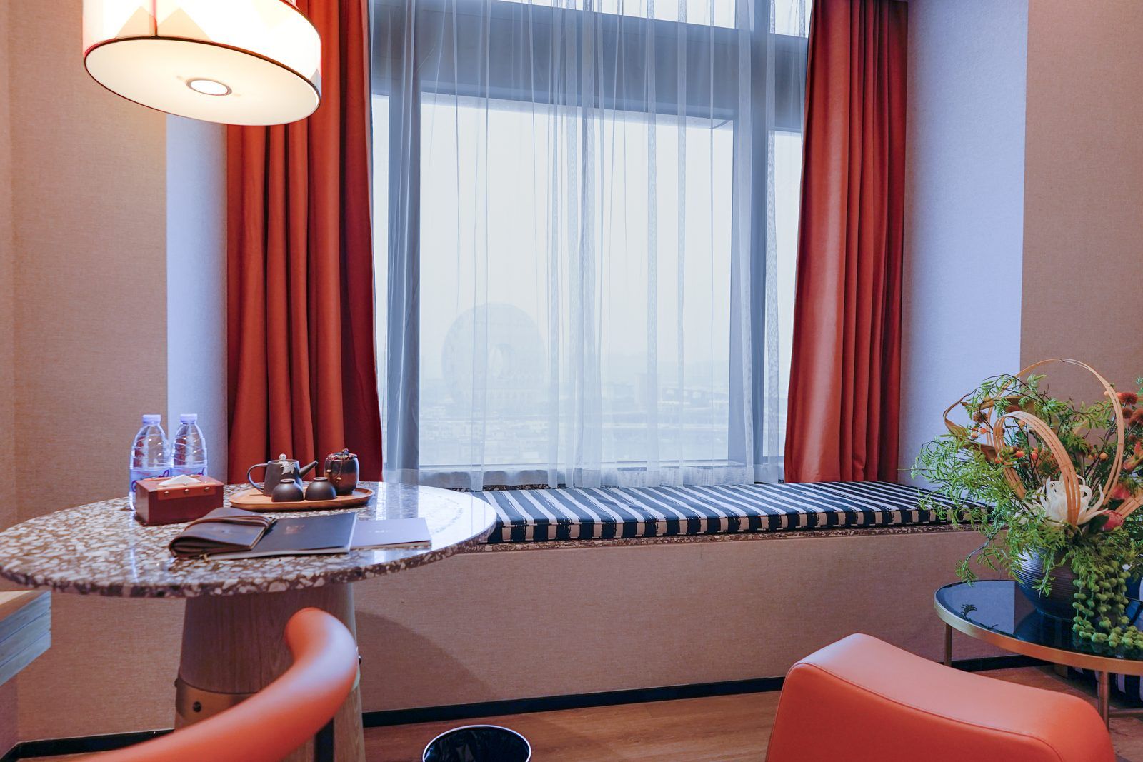 A hotel room with a round table, a bench, a sofa, a window with sheer curtains, and a potted plant.