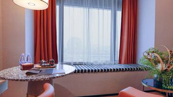 A hotel room with a round table, a bench, a sofa, a window with sheer curtains, and a potted plant.