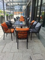 Un patio al aire libre con una mesa de vidrio rodeada de sillas de mimbre con cojines naranja.