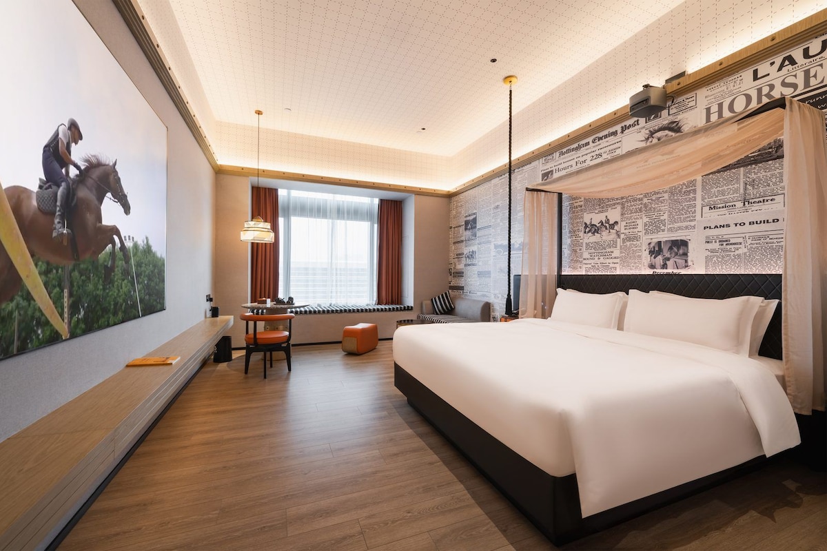 Quarto de hotel com uma cama grande, uma escrivaninha, uma cadeira e janela com cortinas.