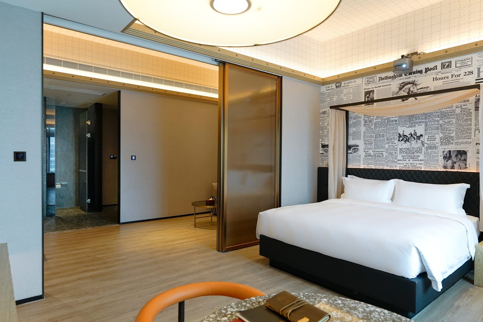 A hotel room with a bed, a desk, and a chair. There is a door leading to another room and a wall with newspaper clippings.