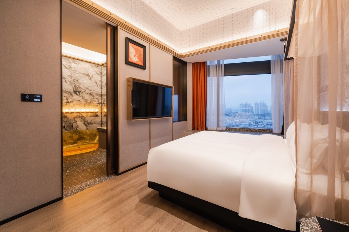 Quarto de hotel com cama, televisão, vista para a cidade e parede de mármore.